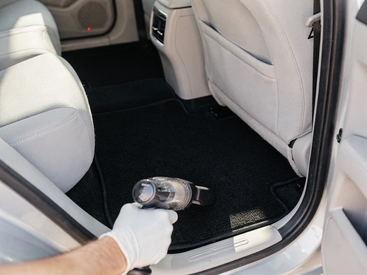 Gloved hand running a small handheld vacuum over the rear floor mat of a clean car with light grey seats